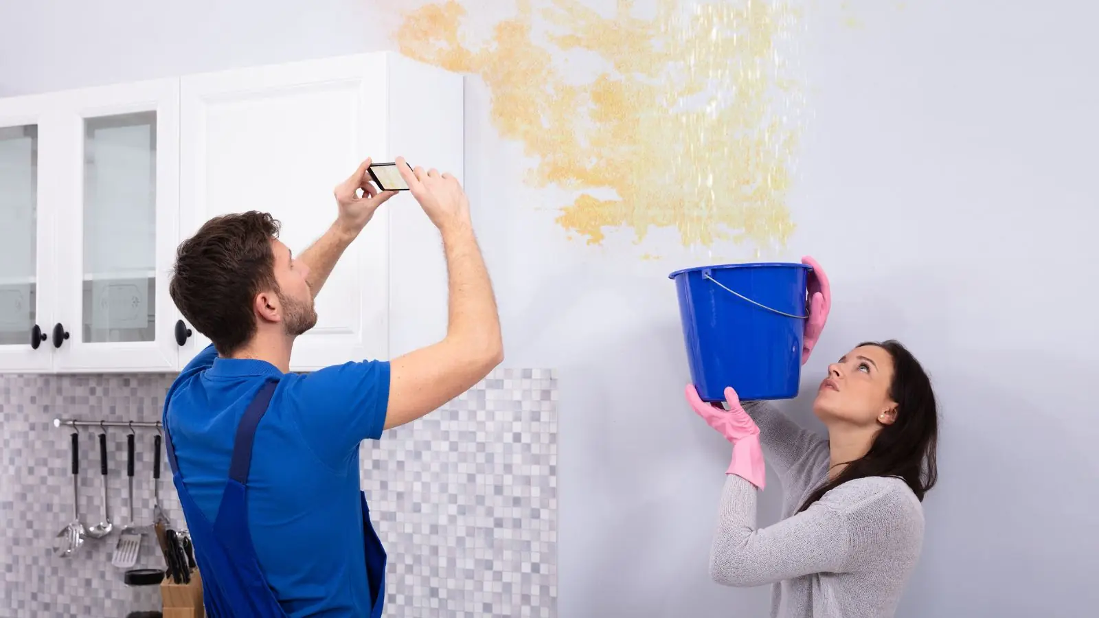 Homeowner photographing a ceiling water stain while another person catches drips in a bucket